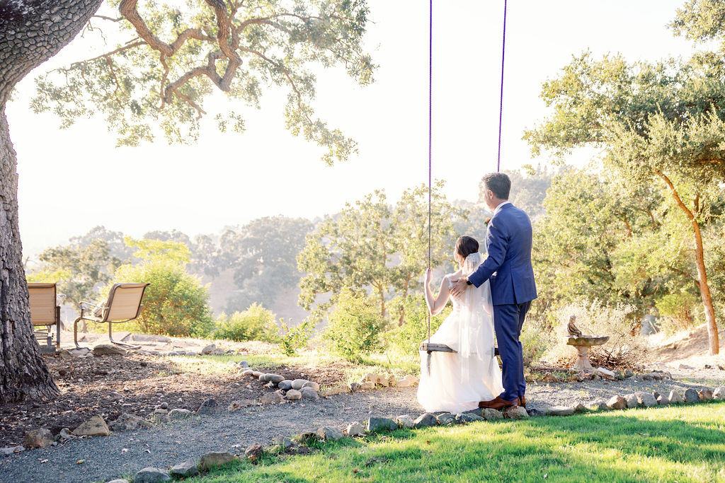 bride sitting on a swing that is hanging from a tree while her groom pushed the swing while standing behind her and the sun sets through the trees