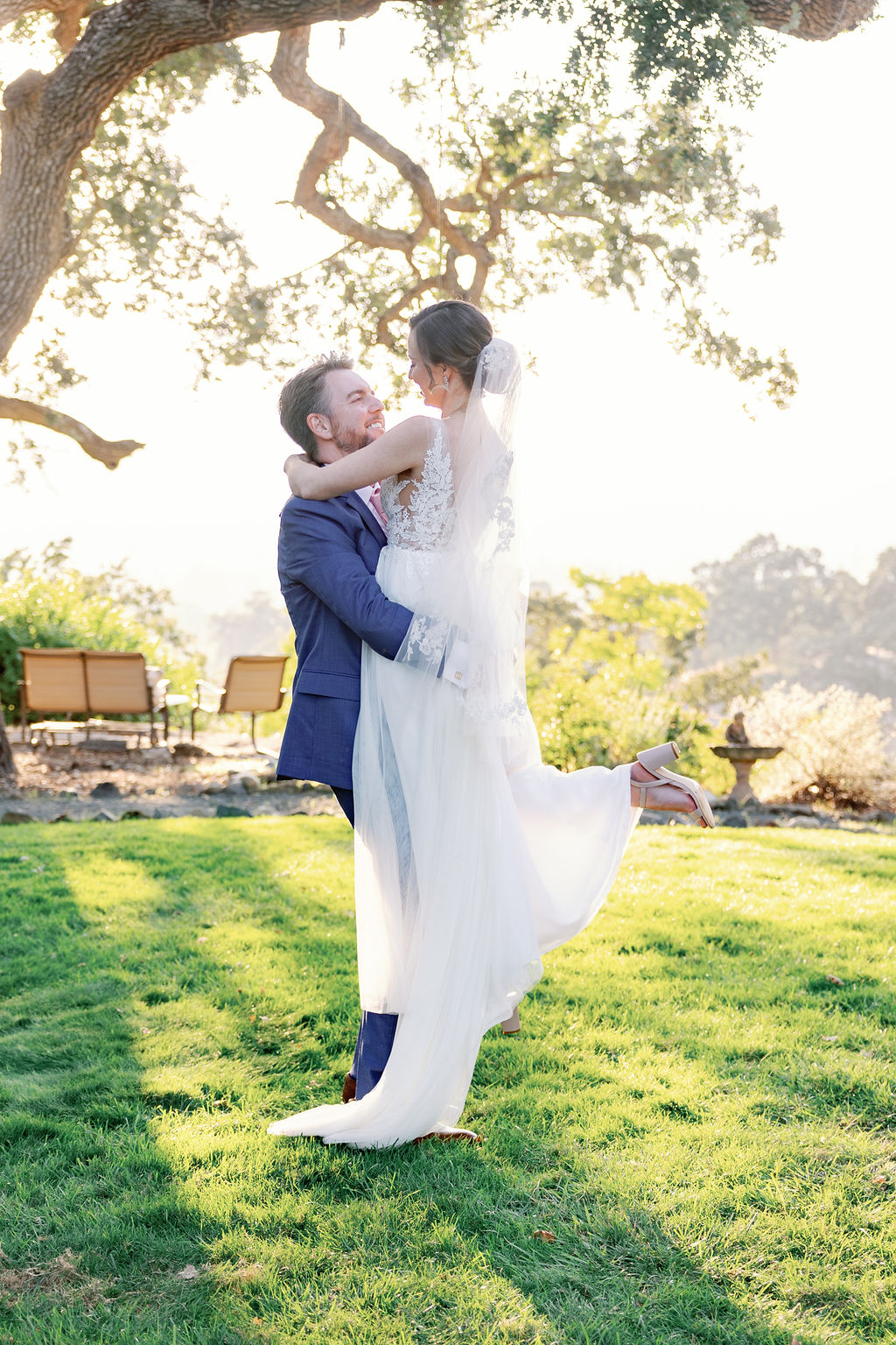 groom picks up his bride and twirls her around while the sun sets through the trees behind them while the shadows cast over them for California wedding