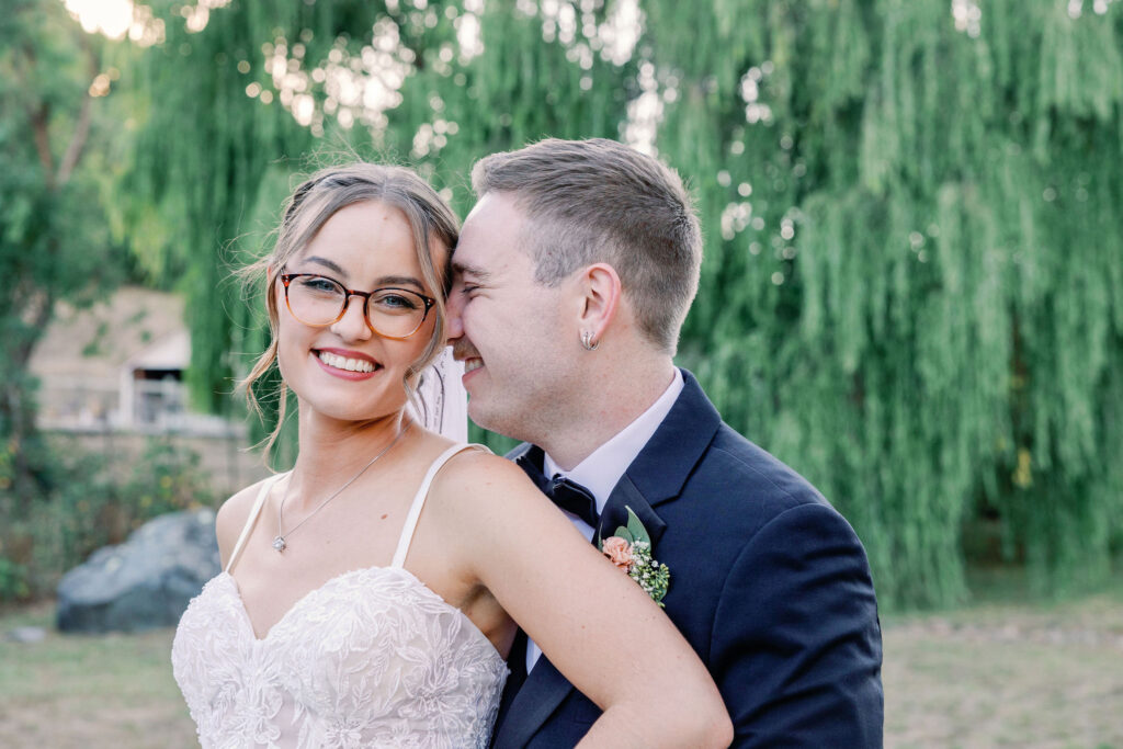 groom holding his bride around her waist and smiling into her neck as she giggles and smiles at the camera held by Bay Area wedding photographers