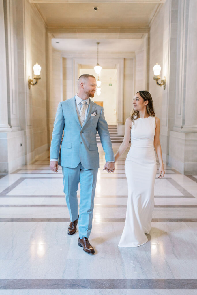 Bay Area wedding photographer captures bride and groom holding hands through city hall and smiling